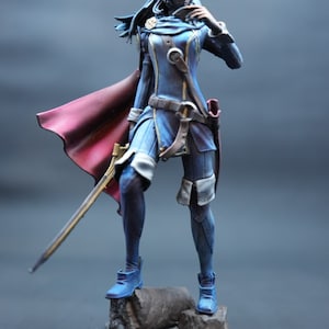 Può includere: Una statuetta dipinta di blu e rosso di un personaggio femminile che indossa un abito blu e oro con un mantello rosso e tiene una spada. La statuetta è in piedi su una base rocciosa.