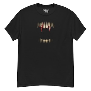 Può includere: T-shirt nero con una grafica della bocca di un vampiro, che mostra denti affilati e sangue che gocciola dalle zanne. La maglietta ha il testo "Creatures + Killers" in alto.