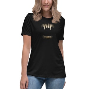 Può includere: T-shirt nero con una grafica stilizzata di denti di vampiro. Il design include denti superiori e inferiori con un effetto luminoso. La maglietta è di vestibilità ampia, adatta per l'abbigliamento casual.