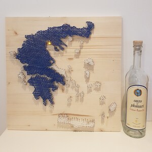 Könnte beinhalten: Ein Wandkunstwerk aus Holz mit einer String-Art-Karte von Griechenland in marineblauem Faden und einem kleinen Licht. Daneben steht eine leere Glasflasche mit dem Etikett "Ouzo of Plomari". Das Kunstwerk befindet sich auf einer hellen Holzplatte.