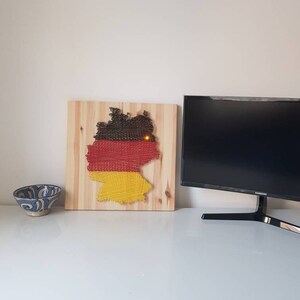 May include: A wooden wall hanging depicting the map of Germany, crafted with brown, red, and yellow string. The artwork is mounted on a light-colored wooden board. A small light is embedded in the center of the map. A decorative bowl and a computer monitor are also visible.
