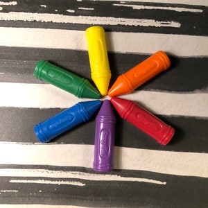 May include: Six crayons arranged in a star shape on a black and white striped surface. The crayons are red, orange, yellow, green, blue, and purple.