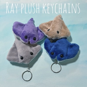 Puede incluir: Cuatro llaveros de peluche con forma de raya en tonos morado, gris, beige y azul. Cada llavero tiene un pequeño ojo negro redondo y un llavero plateado. El texto "RAY PLUSH KEYCHAINS" está escrito en blanco en la parte superior.