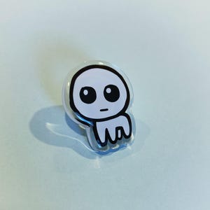 May include: A clear, rounded pin with a cartoon ghost design. The ghost has a white body, large black eyes, and a small black mouth. The pin is set against a light blue background.