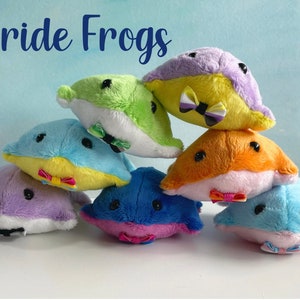 May include: A selection of colourful plush frog toys, each with a bow tie, stacked in a pyramid. The frogs are in shades of blue, green, orange, and purple. The text "Pride Frogs" is visible at the top.