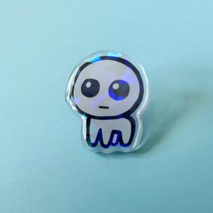 May include: A clear, iridescent pin featuring a cartoon-style character with large, black eyes and a small mouth. The character has a rounded head and a body with a wavy, stylized design. The pin is set against a light blue background.