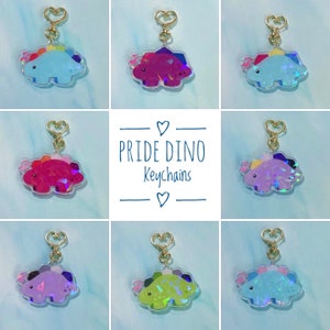 Pode incluir: Uma coleção de chaveiros Pride Dino em várias cores, incluindo azul, rosa, roxo e verde. Cada chaveiro apresenta um design de dinossauro com um fecho dourado em forma de coração. O texto "Pride Dino Keychains" é exibido.