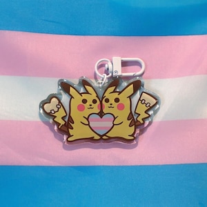 Puede incluir: Un llavero acrílico transparente con dos personajes Pikachu amarillos abrazándose, con un corazón con los colores de la bandera transgénero. El llavero tiene un cierre plateado y está sobre un fondo de la bandera del orgullo transgénero.
