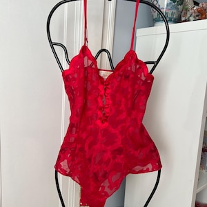 May include: A red, one-piece lingerie garment with a floral pattern, hanging on a black metal hanger. The lingerie has thin straps and a scalloped neckline. The garment is made of a sheer, lightweight fabric.