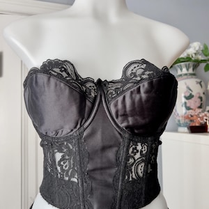 May include: Black satin corset with lace trim, displayed on a white mannequin. The corset features a strapless design with a sweetheart neckline, underwire cups, and delicate lace detailing along the edges and center panel. The garment is a lingerie item.