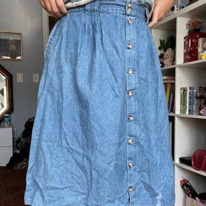 May include: Light blue denim skirt with a button-down front. The skirt has a high waist and a knee-length hemline. The buttons are a light brown color. The skirt is made of a medium-weight denim fabric.