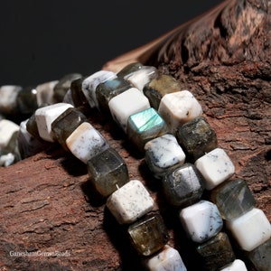 Puede incluir: Una hebra de cuentas de piedras preciosas blancas y grises con acentos iridiscentes. Las cuentas son de forma cúbica y tienen un patrón moteado. Las cuentas están dispuestas en línea sobre una superficie de madera marrón.