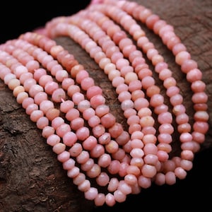 May include: Strands of faceted pink opal beads. The beads are a soft pink color and have a slightly irregular shape. The beads are strung together, creating a necklace or bracelet. The background is a dark, textured surface.