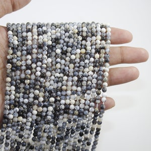 May include: A strand of white and black speckled round beads. The beads are approximately 4mm in diameter and are strung on a clear elastic cord.