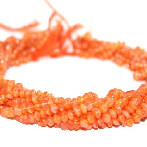 Könnte beinhalten: Eine Strähne orangefarbener Karneolperlen, jede Perle hat eine einzigartige, unregelmäßige Form. Die Perlen sind zu einer langen Halskette aufgereiht.