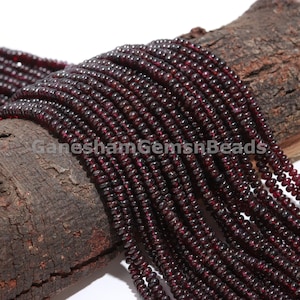 Puede incluir: Primer plano de múltiples hebras de pequeñas cuentas redondas de granate rojo oscuro. Las cuentas están ensartadas, creando una apariencia fluida y texturizada. Las cuentas se exhiben sobre un trozo de madera marrón oscuro. El texto "GaneshamGemsnBeads" es visible.