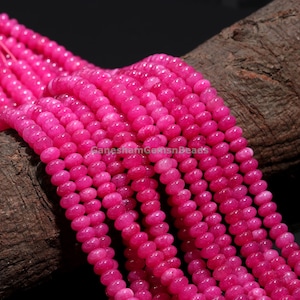 Könnte beinhalten: Eine Strähne aus leuchtend rosa runden Perlen aus Jade. Die Perlen sind glatt und poliert und auf einem durchsichtigen Faden aufgefädelt. Die Perlen sind in einer lockeren Spirale angeordnet und liegen auf einem Stück braunem Holz.