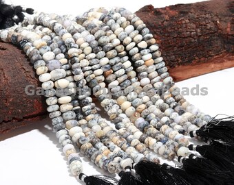 May include: A strand of white and gray opal beads with black tassels. The beads are round and have a speckled appearance. The strand is lying on a piece of wood.