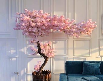 May include: A decorative artificial cherry blossom tree with pink flowers and a brown trunk. The tree is in a wooden pot and placed in front of a white wall. A teal sofa with a matching pillow is in the background.