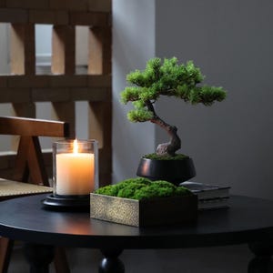 K&ouml;nnte beinhalten: Ein kleiner k&uuml;nstlicher Bonsai mit gr&uuml;nem Blattwerk und braunem Stamm in einer schwarzen Schale, ein quadratischer Beh&auml;lter gef&uuml;llt mit gr&uuml;nem Moos und eine wei&szlig;e Stumpenkerze in einem klaren Glashalter, arrangiert auf einem runden schwarzen Tisch.