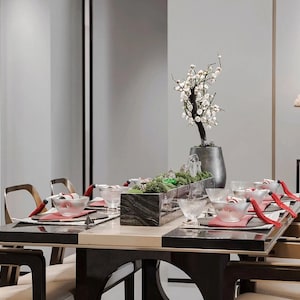 May include: A dining table set for a meal, featuring a black and beige tabletop, surrounded by wooden chairs. The table is set with bowls, plates, and decorative elements, including a floral arrangement and a mountain-shaped sculpture. The room has a minimalist design.