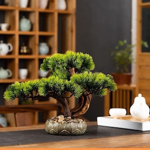 May include: An artificial bonsai tree with green foliage and a brown trunk and branches, displayed in a grey ceramic pot with a lotus flower design. The tree is positioned on a wooden table with a brown placemat.