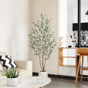 K&ouml;nnte beinhalten: Ein k&uuml;nstlicher Olivenbaum in einem wei&szlig;en Topf steht in einem modernen Wohnzimmer. Der Baum hat gr&uuml;ne Bl&auml;tter und kleine schwarze Oliven. Im Hintergrund befindet sich ein beiges Sofa mit einem schwarz-wei&szlig;en Zickzack-Kissen. Eine kleine Topfpflanze steht auf einem Marmor-Couchtisch.