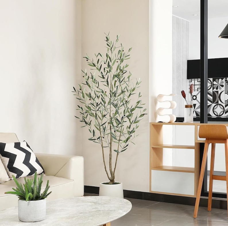 K&ouml;nnte beinhalten: Ein k&uuml;nstlicher Olivenbaum in einem wei&szlig;en Topf steht in einem modernen Wohnzimmer. Der Baum hat gr&uuml;ne Bl&auml;tter und kleine schwarze Oliven. Im Hintergrund befindet sich ein beiges Sofa mit einem schwarz-wei&szlig;en Zickzack-Kissen. Eine kleine Topfpflanze steht auf einem Marmor-Couchtisch.