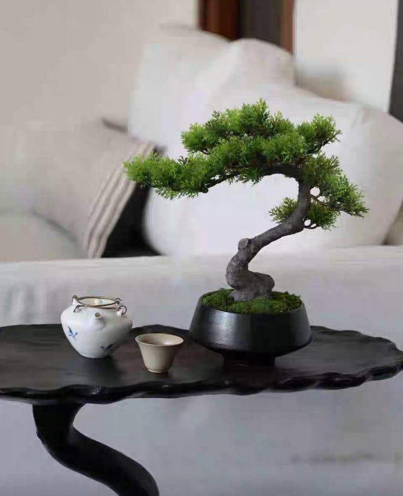 K&ouml;nnte beinhalten: Ein kleiner k&uuml;nstlicher Bonsai-Baum mit leuchtend gr&uuml;nem Laub und einem geschwungenen dunklen Stamm, gepflanzt in einem runden schwarzen Keramiktopf. Neben dem Bonsai stehen eine kleine wei&szlig;e Keramik-Teekanne mit blauen Blumenmustern und eine beige Teetasse.