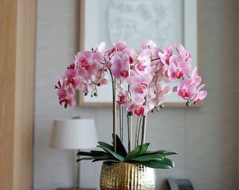以下が含まれることがあります： 金色のハンマードメタルの花瓶にピンクの蘭の花束。蘭の花は円形に配置され、緑の葉がついています。花瓶は木製のテーブルの上に置かれています。