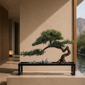 May include: A bonsai tree with green leaves and a curved trunk in a black rectangular planter. The planter holds small, grey rock formations. The bonsai is displayed on a beige platform, with a mountain and water feature in the background.