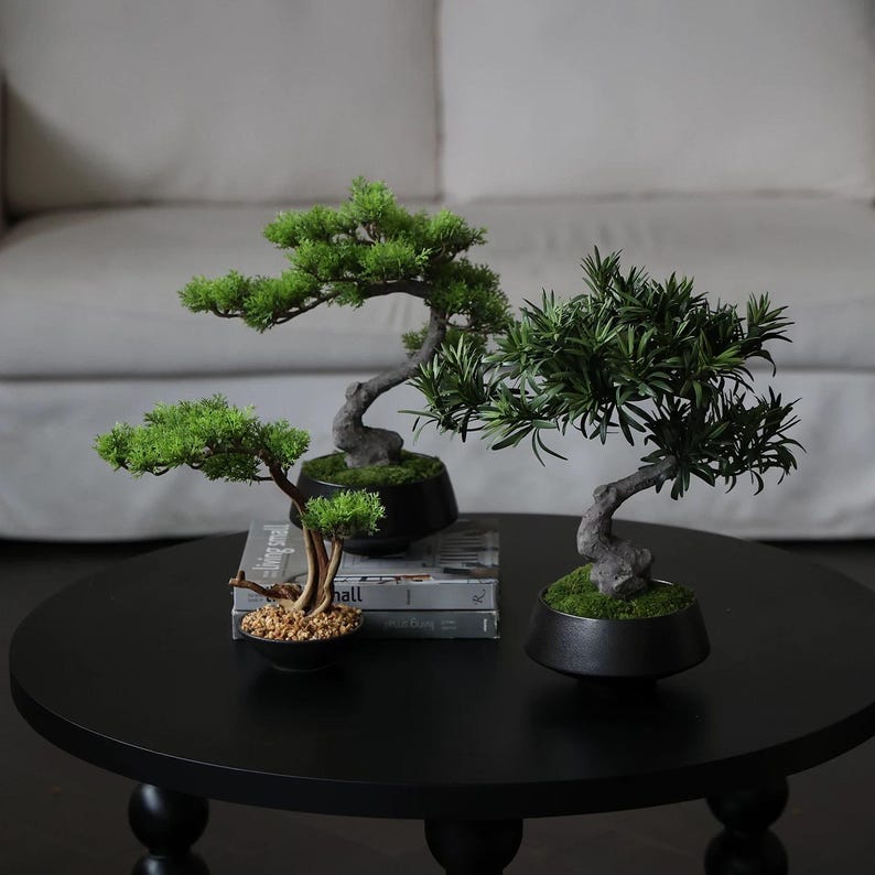 K&ouml;nnte beinhalten: Zwei k&uuml;nstliche Bonsai-B&auml;ume mit gr&uuml;nem Blattwerk und strukturierten grauen St&auml;mmen, jeweils in einer mattschwarzen Schale. Ein Baum hat einen geschwungenen Stamm mit moosiger Erde, der andere eine kleinere Basis mit hellen Kieselsteinen.