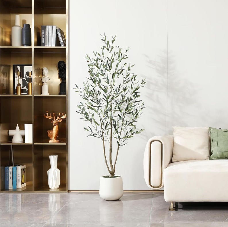 K&ouml;nnte beinhalten: Ein k&uuml;nstlicher Olivenbaum mit gr&uuml;nen Bl&auml;ttern und einem braunen Stamm in einem wei&szlig;en Topf. Der Baum steht vor einer wei&szlig;en Wand, neben einem cremefarbenen Sofa mit gr&uuml;nen und cremefarbenen Kissen. Ein goldfarbenes Regal ist links zu sehen.