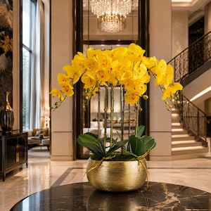 Peut inclure: Un arrangement vibrant de fleurs d'orchidées jaune vif avec des feuilles vertes, soutenu par de fines tiges verticales, planté dans un bol métallique rond et texturé de couleur dorée posé sur une surface circulaire polie.