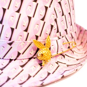 Puede incluir: Sombrero fedora rosa con un patrón repetido de siluetas de conejos blancos sobre óvalos rosas y rectángulos negros. Un broche de conejo dorado con cierre de imperdible está adjunto.