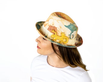 May include: A floral print fedora hat with a cream-colored ribbon and a gold-colored lizard-shaped embellishment. The hat features a mix of colors, including beige, green, and brown. The hat is a stylish accessory.
