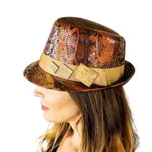 Peut inclure: Un chapeau fedora avec un motif peau de serpent dans les tons marron, orange et or. Le chapeau est orné d'un ruban beige avec trois ornements carrés dorés. Le bord est également beige. Le chapeau est sur fond blanc.