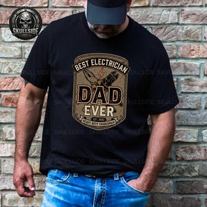 May include: A black t-shirt featuring a graphic design with the text BEST ELECTRICIAN DAD EVER, EST. 2023, and HEAVY DUTY TRADESMAN. The design includes illustrations of pliers and wire cutters with a lightning bolt icon in a tan-colored badge.