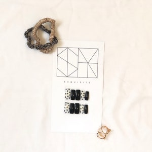 Könnte beinhalten: Draufsicht auf einer weißen Oberfläche mit schwarzen Kunstnägeln mit Schmetterlingsakzenten, einer Karte mit dem Wort "EXQUISITE", zwei Haargummis und einem Paar goldener Ringe. Die Nägel sind rechteckig.
