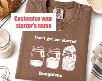 May include: An espresso-colored t-shirt featuring a white graphic of three glass jars with tags, the text "Don't get me started" and "Doughlene," and a "Customize your starter's name" box. The shirt has a Comfort Colors label at the inner neckline.