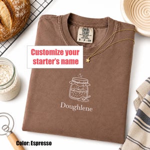 May include: An espresso-colored t-shirt featuring a white line-art graphic of a sourdough starter jar with a spoon and the text "Doughlene" below it. The shirt has a Comfort Colors label at the neck and a gold star pendant necklace resting on it.