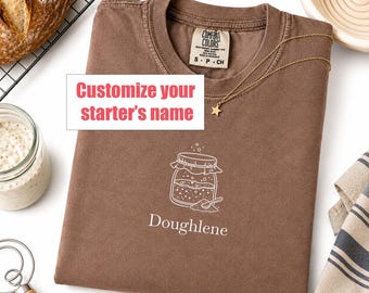 May include: An espresso-colored t-shirt featuring a white line-art graphic of a sourdough starter jar with a spoon and the text "Doughlene" below it. The shirt has a Comfort Colors label at the neck and a gold star pendant necklace resting on it.