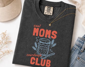 May include: A graphite-colored short-sleeve t-shirt featuring a graphic print with the text "cool MOMS sourdough CLUB". The design includes a central jar illustration surrounded by wheat stalks and bubbles, rendered in coral and light blue ink colors.