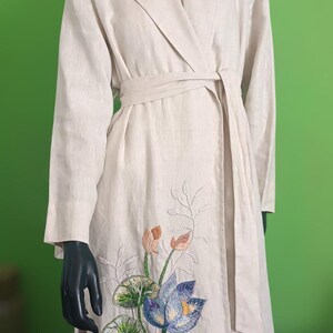 Könnte beinhalten: Ein langer, beiger Leinen-Wickelmantel mit einem floralen Stickmuster mit blauen, grünen und orangefarbenen Blumen. Der Mantel hat einen selbstbindenden Gürtel und eine lange, fließende Silhouette.