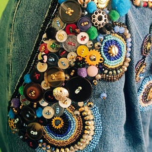 Könnte beinhalten: Hellblaue Jeans mit aufwendigen Verzierungen. Die Jeans haben einen Patchwork aus bunten Knöpfen, Perlen und dekorativen Elementen. Das Design umfasst Knöpfe, Perlen und kleine Pompons in verschiedenen Größen und Farben.