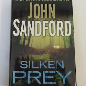 May include: A book cover with a dark green and black background. The title "Silken Prey" is in large, blue letters. The author's name, "John Sandford", is in white letters above the title. The book is a novel.