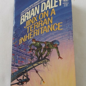 May include: A science fiction paperback book cover with a blue background and a yellow title, "Brian Daley Jinx on a Terran Inheritance". The cover features an illustration of two people in spacesuits on a metal platform.