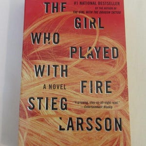 Può includere: Una copertina di libro rossa con il titolo "The Girl Who Played With Fire" di Stieg Larsson. La copertina presenta un primo piano dei capelli biondi di una donna.