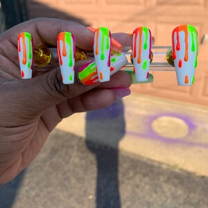 May include: Set of five white press-on nails with a drip design. The nails are painted with bright neon green, orange, and pink colours.