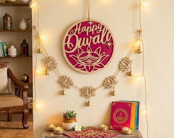 May include: A circular wooden wall sign with the text Happy Diwali in script, featuring a cutout diya lamp design over a magenta background. A string of warm white lights and wooden lotus flower ornaments with small brass bells hang below.